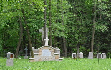 pomnik z krzyżem obok mniejsze nagrobki, drzewa, trawa