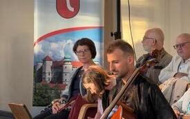 mężczyzna gra na wiolonczeli, uczestnicy koncertu, w tle roll-up