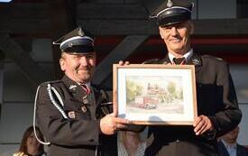 dw&oacute;ch strażak&oacute;w z pamiątkowym obrazem przedstawiającym samoch&oacute;d OSP na tle kościoła
