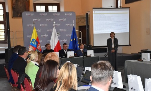 przemawia burmistrz, radni powiatowi siedzą przy stole, flagi banery w tle
